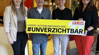 Four people stand together holding a yellow banner that reads Unsere Unternehmer:innen sind #unverzichtbar. They are smiling and posing for a photo in a restaurant.