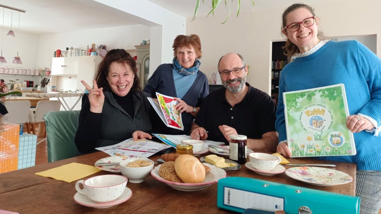 Five people sit at a table with bread, coffee, and snacks. They smile, holding a magazine. A binder is on the table.