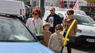 A family with two children stands in front of a blue car, two adults holding a balloon, and two kids in front, all wearing sunglasses. Behind them, a man and a woman, and flags.