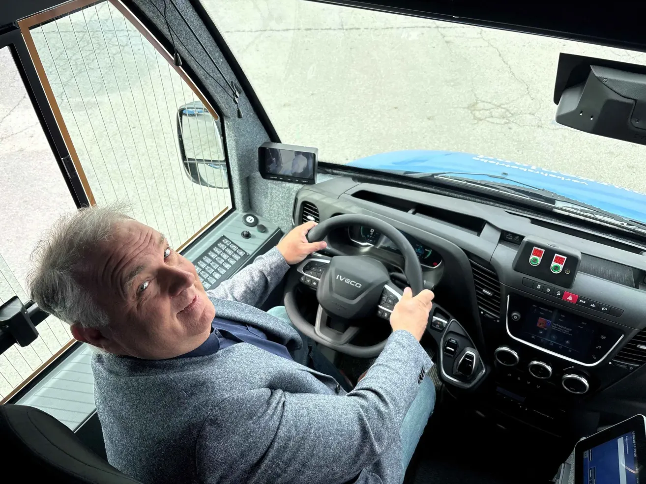 An older man sits in the driver's seat of a blue vehicle, holding the steering wheel. The interior is modern with various controls and a rear-view mirror.