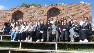 A group of people sits on wooden benches in front of a brick wall, smiling for a photo. They are all dressed casually, with some wearing sunglasses.
