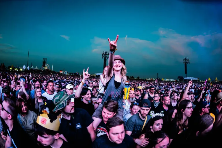 A large crowd of people at an outdoor concert, some with peace signs, one woman sitting on a man's shoulders, smiling and waving. The sky is clear with a few clouds.