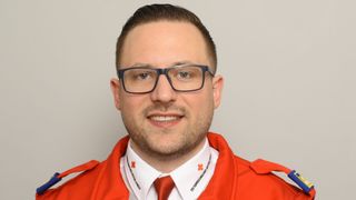 A smiling man in a red jacket with a red cross symbol on his collar. He wears glasses and a red tie. The background is plain.