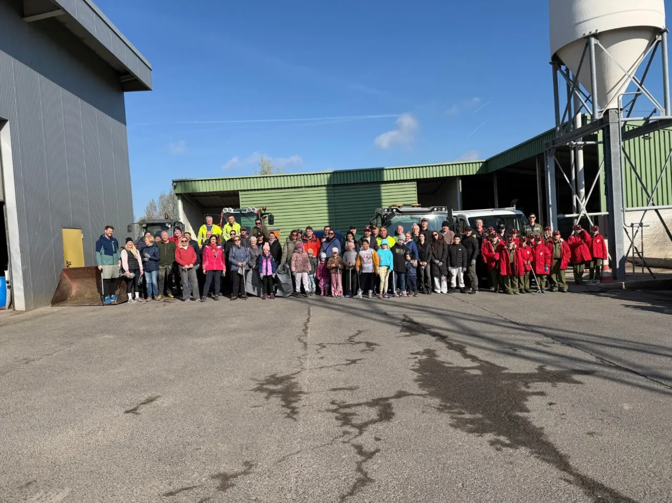 A group of people stands in front of a building, some in red uniforms, others in jackets. Behind them, trucks are parked. The sky is clear.