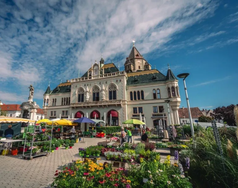 A large ornate building with a clock tower stands in the center of a market square. It is surrounded by vibrant flowers and plants. People walk among the stalls. The sky is partly cloudy.