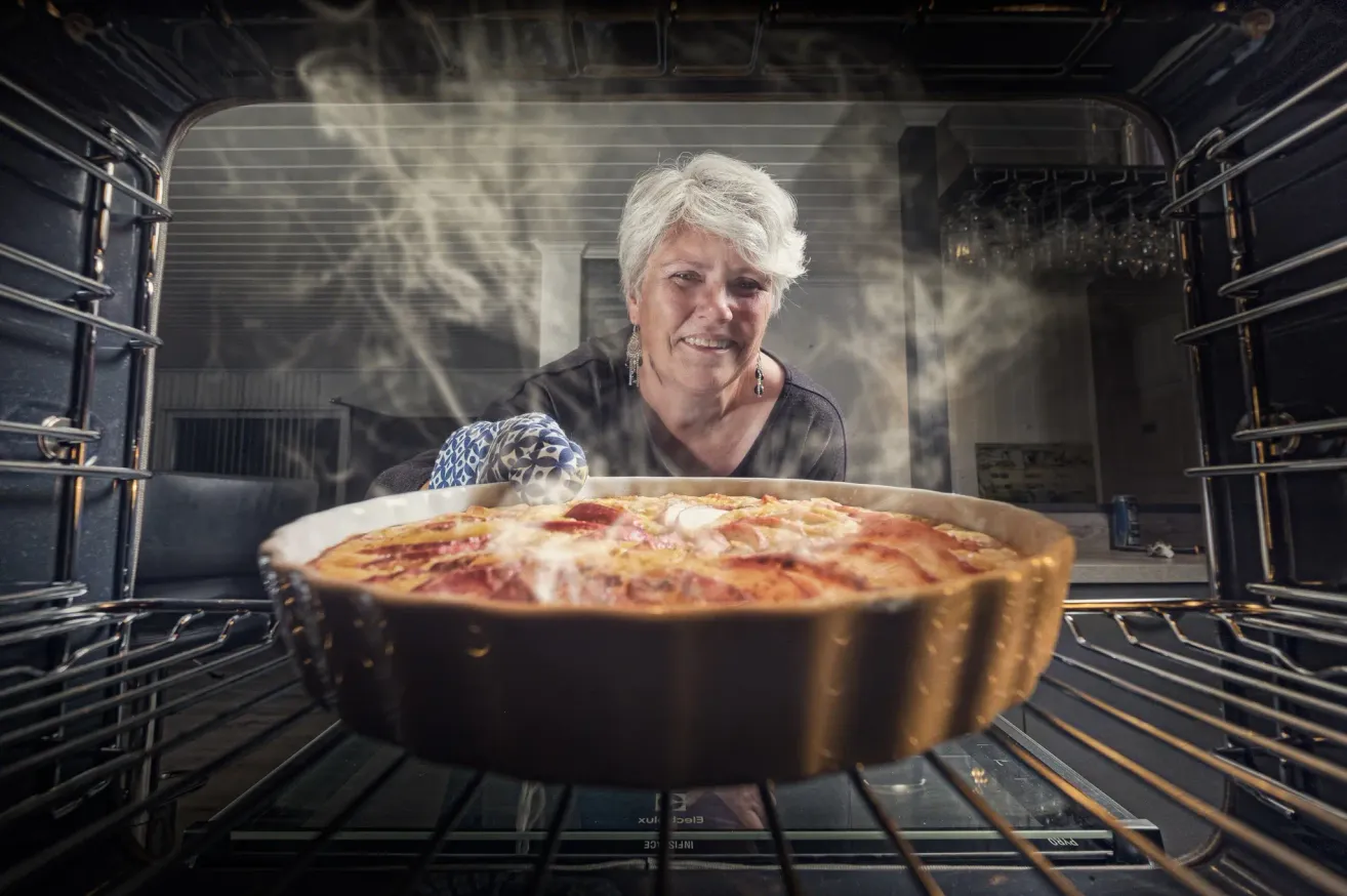 A smiling woman wearing gloves holds a steaming pie in an open oven. The oven is filled with smoke.