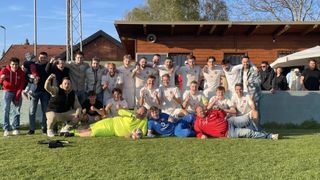 A soccer team poses for a photo on a field with a building in the background. They are wearing white uniforms and some are in casual clothing.
