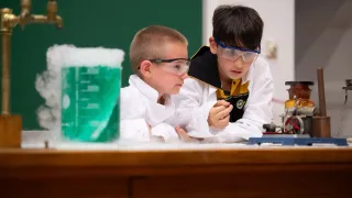 Two young boys wearing goggles and lab coats are conducting a science experiment. One boy is looking into a green beaker while the other boy is holding a small object.