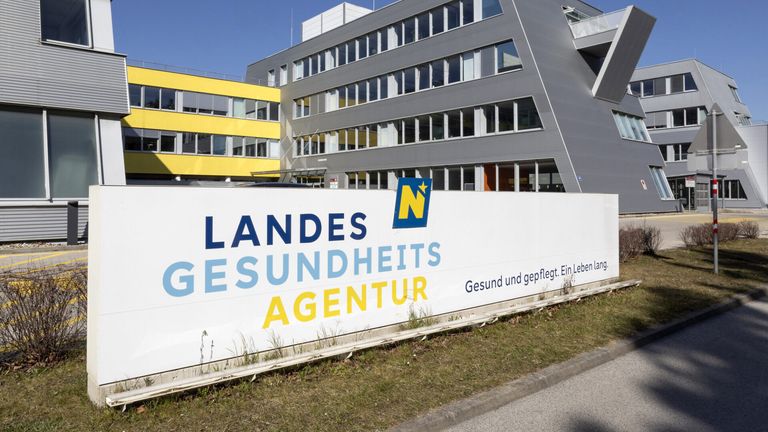 A large sign for the Landesgesundheitsagentur is displayed in front of a modern building with glass windows. The sign is white with blue and yellow text.