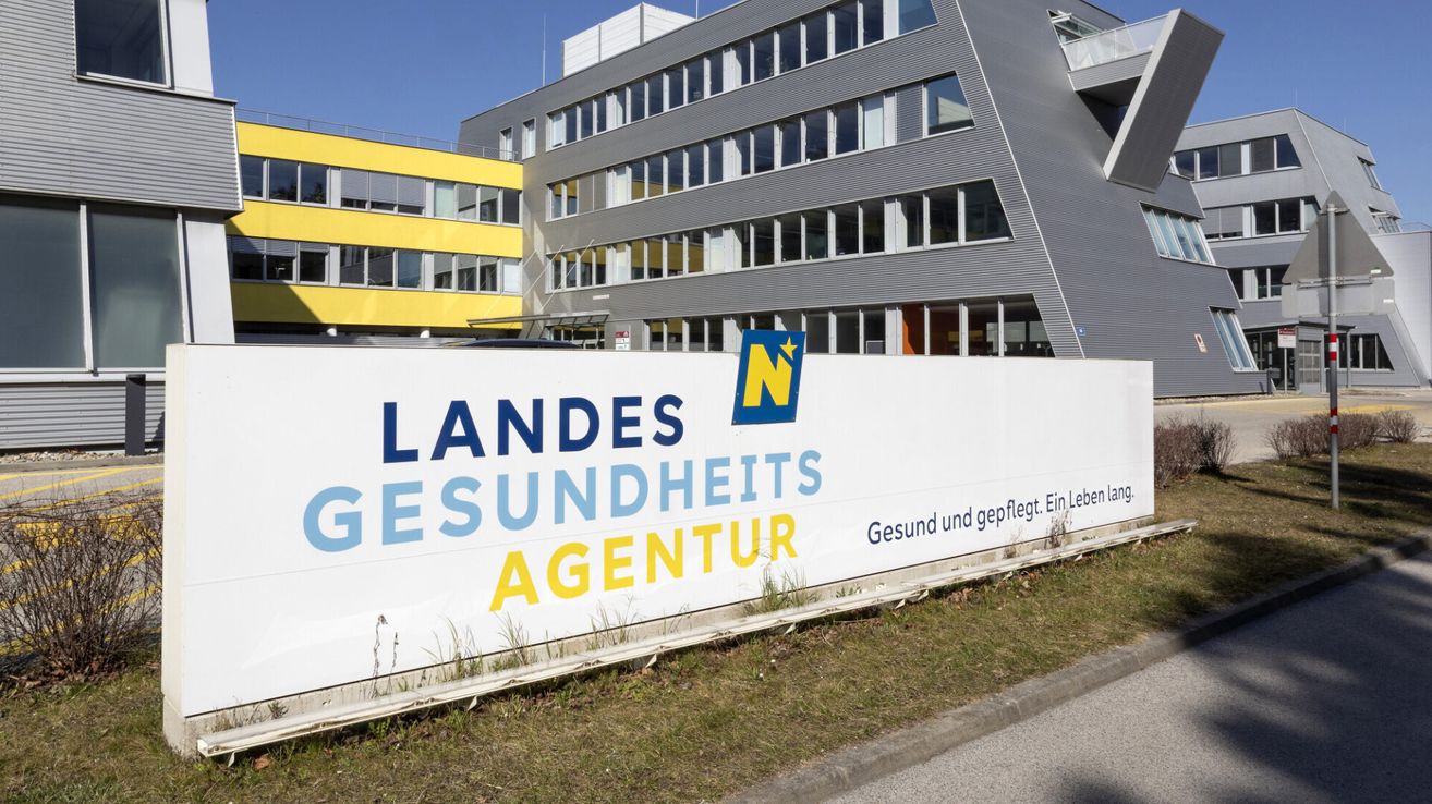 A large sign for the Landesgesundheitsagentur is displayed in front of a modern building with glass windows. The sign is white with blue and yellow text.