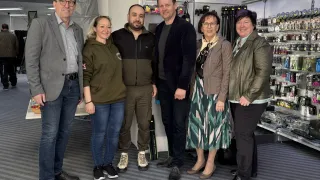 Six individuals, three men and three women, stand together in a room with a striped carpet. They are smiling and posing for a photo. Behind them, there is a shelf filled with various items.