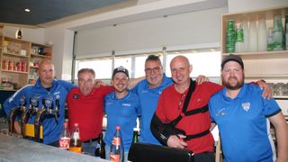 Five men wearing blue and red shirts with a USCP logo, standing together and smiling. One has a black arm brace. They are standing by a bar with various bottles and a black monitor. Behind them are glass windows.