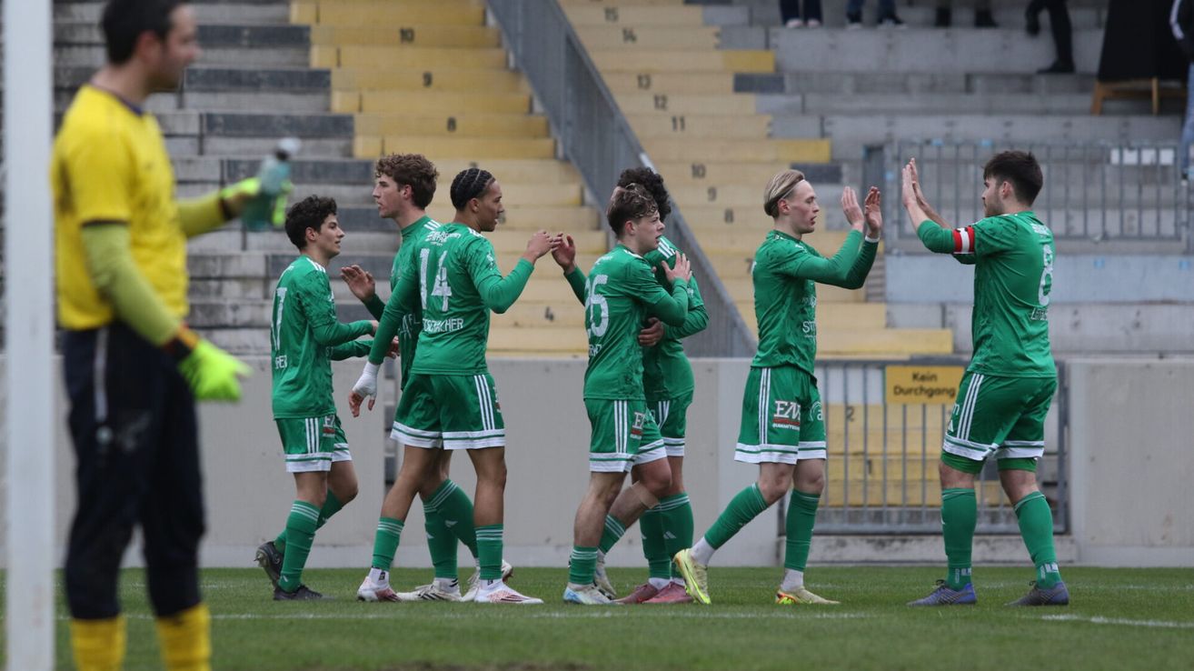 A soccer team in green uniforms celebrates on the field. Numbers and logos are visible on their jerseys. An empty stand with yellow steps is behind them.