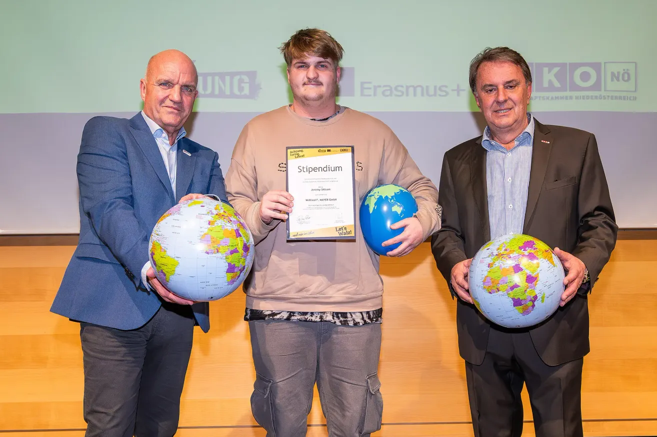 Three men stand together holding globes. The man in the middle holds a certificate, smiling. The man on the left wears a blue suit, and the man on the right wears a brown suit. The background is green with text.