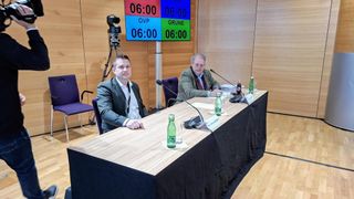 Two men sit at a table with microphones. One wears a suit, the other a sweater. A digital timer displays 06:00. A tripod and camera are behind them.