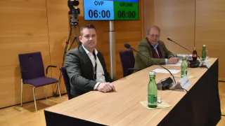 Two men sit at a table with microphones, one wearing a suit and the other a jacket. Behind them, a digital timer shows 06:00, and a camera tripod is nearby.