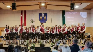 A band performs on stage with musical instruments in front of an audience. They wear traditional attire. A large screen displays text and musical notes.