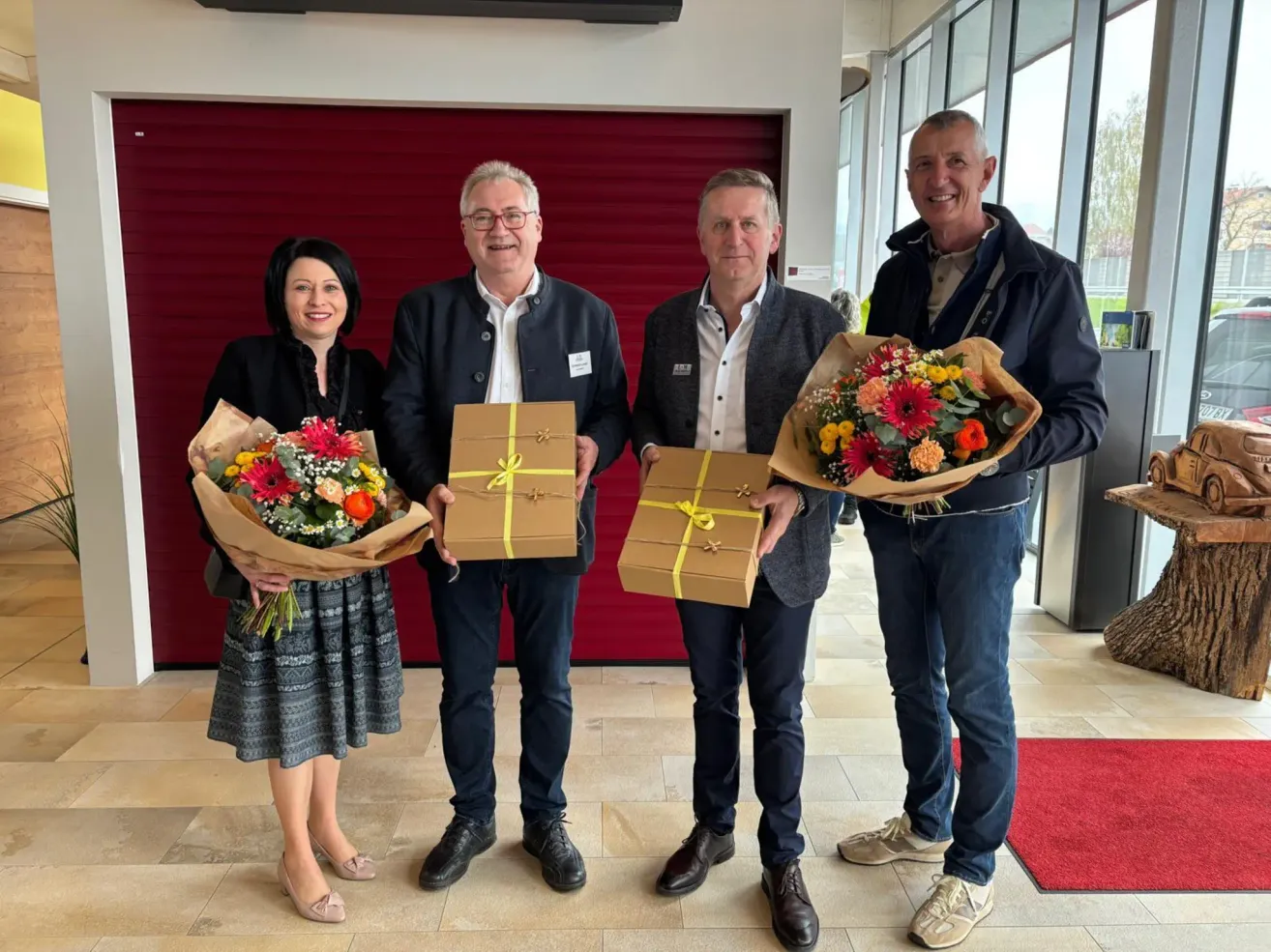 Four people, three men and one woman, stand together holding flowers and wrapped gifts. They are smiling and appear to be at a celebration.