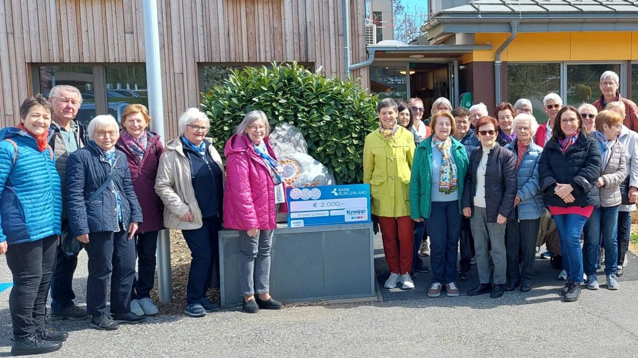A group of women stands outside, smiling, with a donation box marked with 2,000 euros. Behind them is a building with a green plant and a blue sign.