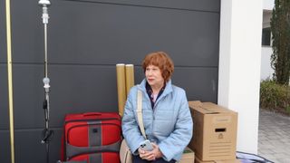 An elderly woman with red hair, wearing a blue jacket, holds a cell phone and stands in front of a wall with luggage and boxes.