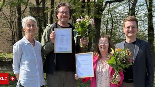 Five people pose for a picture in a park. Two men and three women hold certificates and bouquets. They are smiling and appear to be celebrating.