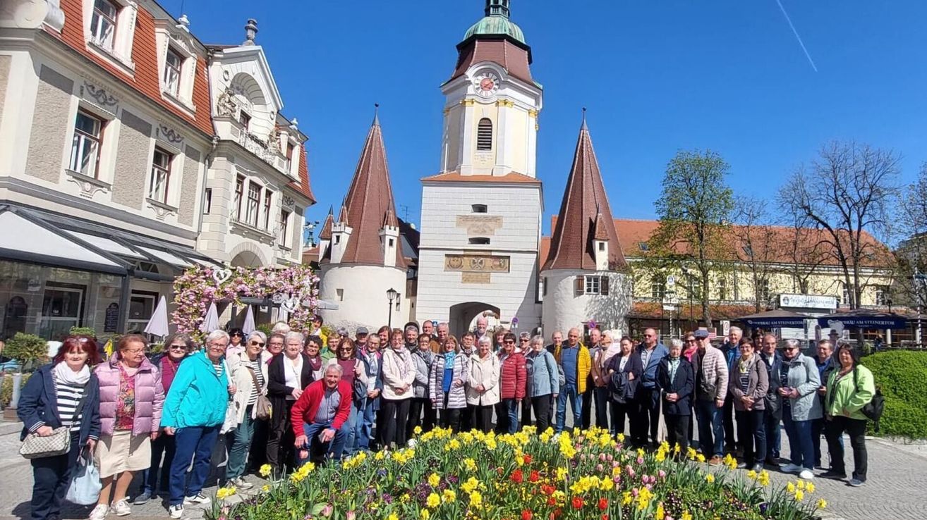 A group of people wearing winter clothes poses in front of a tower with a clock and a garden with yellow and red flowers.