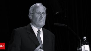 An older man in a suit and tie stands before a microphone, possibly giving a speech. He wears glasses and gestures with his hand.