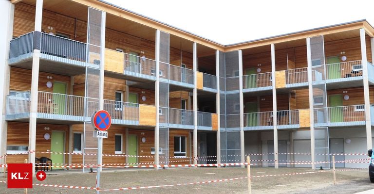 A modern multistory apartment building with wooden exteriors and metal railings, surrounded by a construction fence.