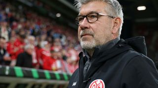 An older man in a black jacket with glasses and a red logo is standing in a stadium with a crowd of people wearing red shirts.