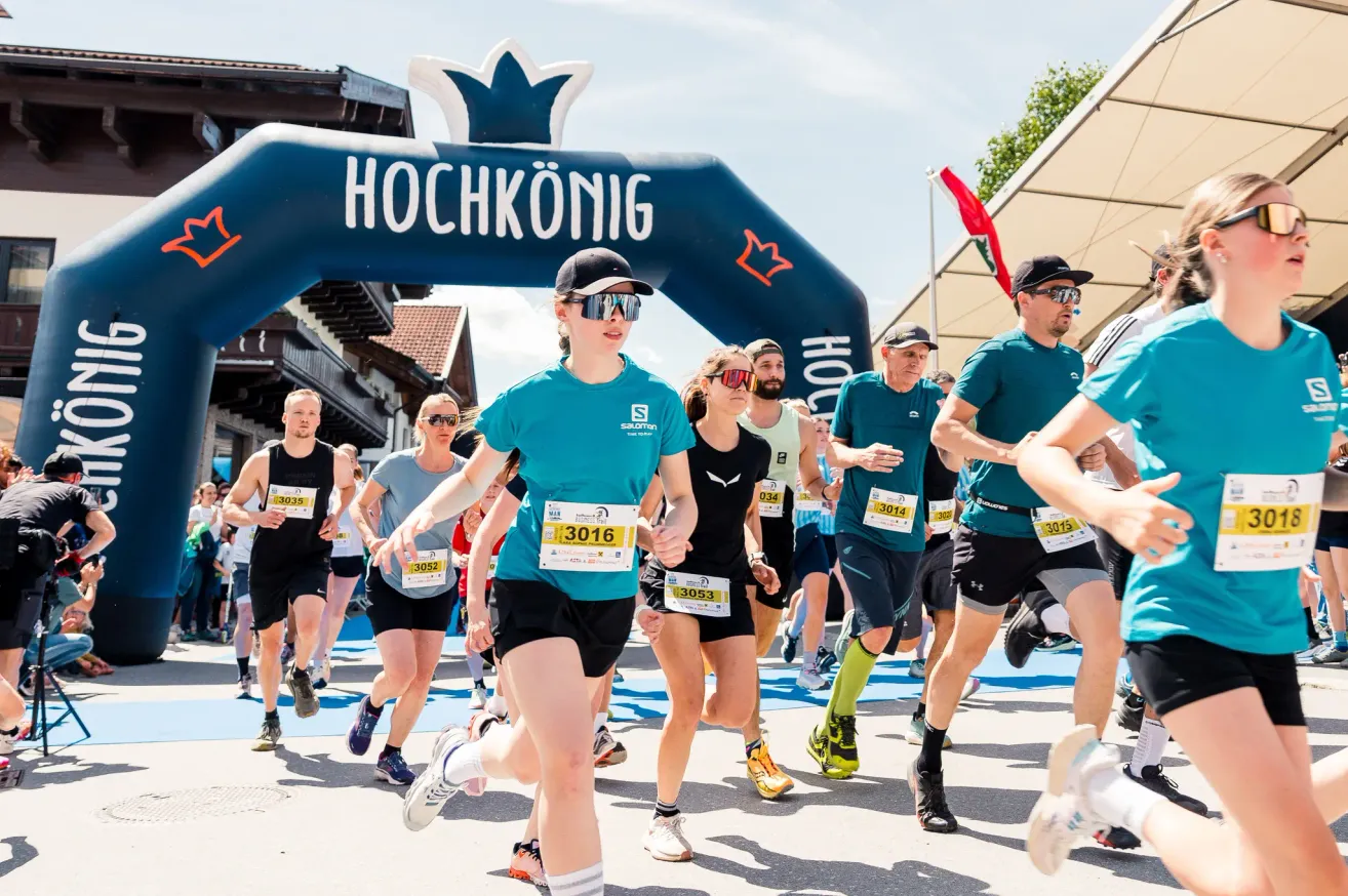 Marathonläufer überqueren die Ziellinie eines Rennens. Ein blauer Bogen mit dem Wort Hochkönig befindet sich über ihnen. Die Läufer tragen Kleidung und Schuhe in verschiedenen Farben. Der Mann vorne trägt eine Brille und einen Hut.
