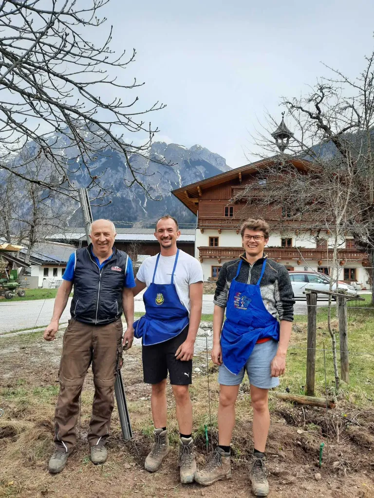 Three men in aprons stand in front of a mountain house with a car parked in front. The man on the left wears a black vest.