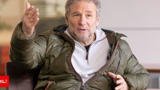 An older man in a green jacket and a white shirt gestures with his hands, likely speaking or explaining something.