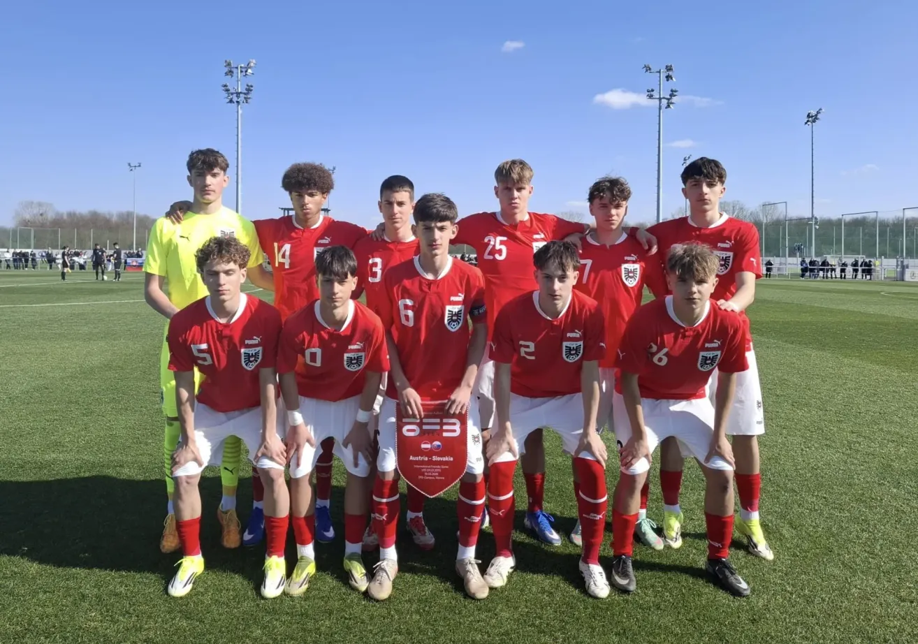 A soccer team in red uniforms stands on a field, posing for a photo with a certificate in the center. The players wear jerseys with numbers and the team's logo. Behind them are two tall light poles.