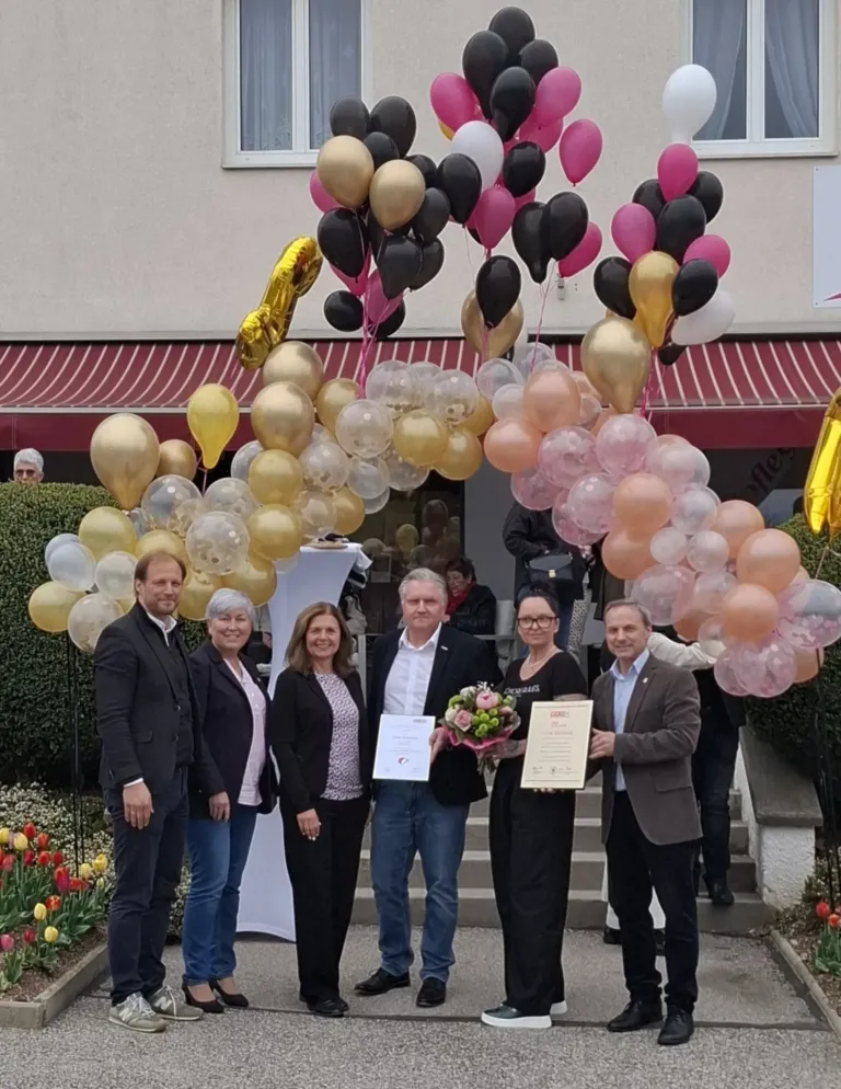 A group of people stand under a balloon arch, holding certificates and flowers. The setting is outdoors, with a building and flowers in the background.