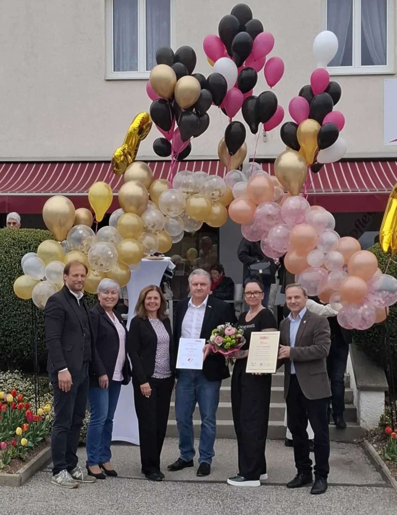 A group of people stand under a balloon arch, holding certificates and flowers. The setting is outdoors, with a building and flowers in the background.
