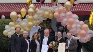 A group of people stand under a balloon arch, holding certificates and flowers. The setting is outdoors, with a building and flowers in the background.