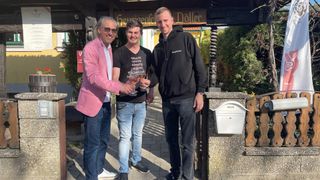 Three men stand outside, smiling. The man on the left holds a wine glass. The middle man wears a black shirt with text. The right man wears a black hoodie. Behind them is a signboard.