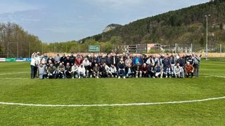 A large group of people are posing for a photo on a soccer field, with a mountain and green sign in the background.