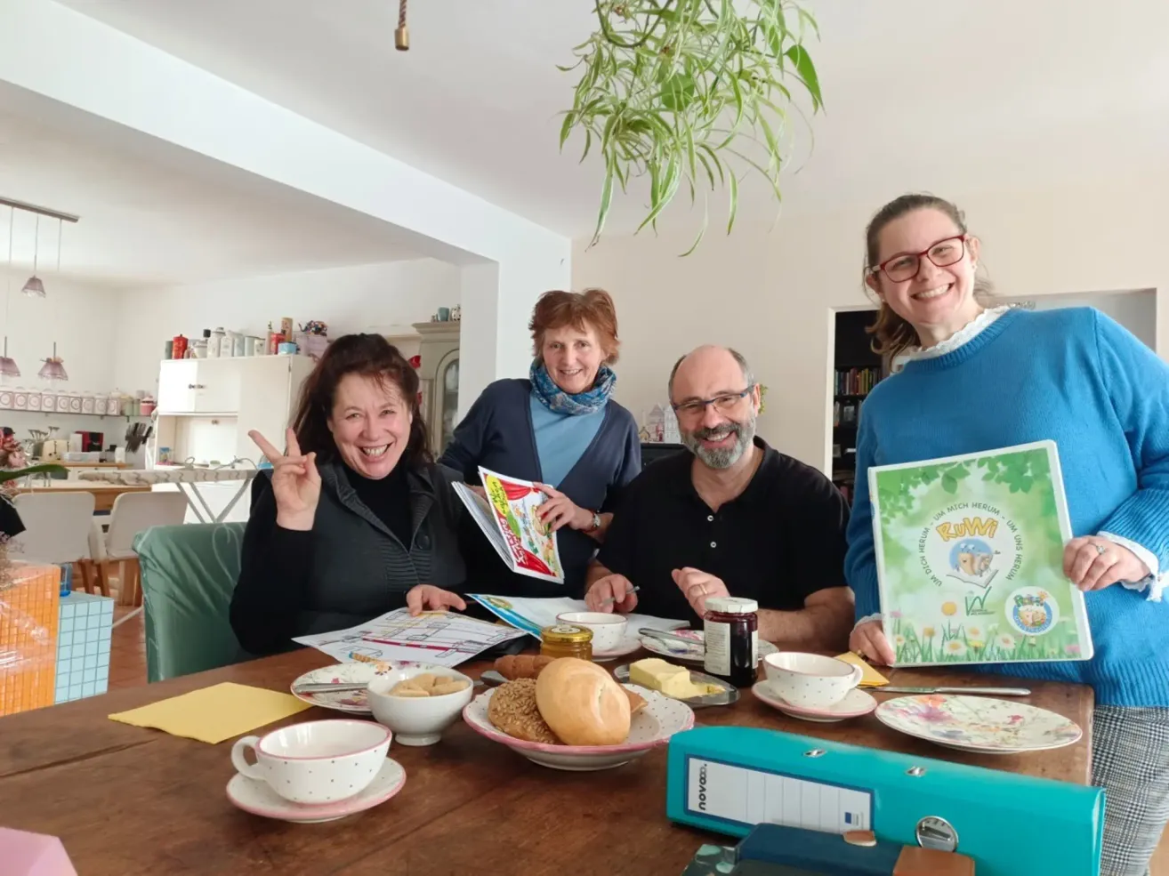 Four people sit at a table, smiling and posing for a picture. A table is set with bread, jams, and tea cups. One person holds a book, while others have magazines. A bookshelf is in the background.