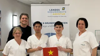 Five medical staff members, two of whom are holding a Vietnamese flag, stand in a room. One is wearing glasses, and the wall behind them has a sign with a logo.