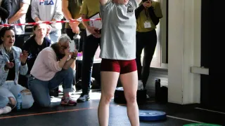 A woman lifts a barbell above her head while others watch, including one man in a Fila hoodie.