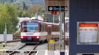 A red Salzburger Lokalbahn train at a station with a digital clock above. The train is on the tracks, and there are signs and trees in the background.