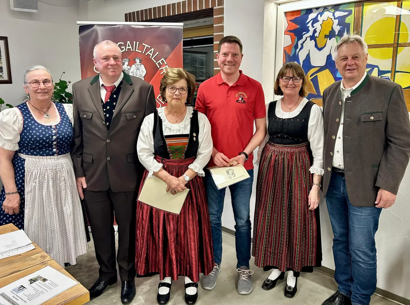 Five people are standing together, smiling, and holding a certificate. The man in the middle is wearing a red shirt and holding a certificate. The woman on the right is wearing glasses.