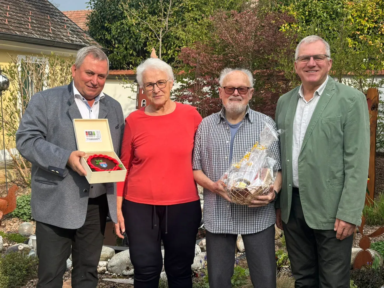 Four people are posing for a photo outdoors. A man on the left holds a framed award. The woman in the middle wears a red shirt. The man on the right holds a basket. Behind them is a house with a garden.