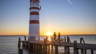 Two people on bikes stand on a dock near a lighthouse as the sun sets on the water.