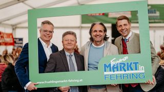 Four men dressed in formal attire stand inside a green frame that reads 'Fruhlingsmarkt in Fehring'. They are all smiling at the camera.