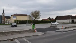 An empty parking lot with buildings in the background. A black car is parked on the left, and a white car is parked on the right. A green trash bin is on the side of the road.