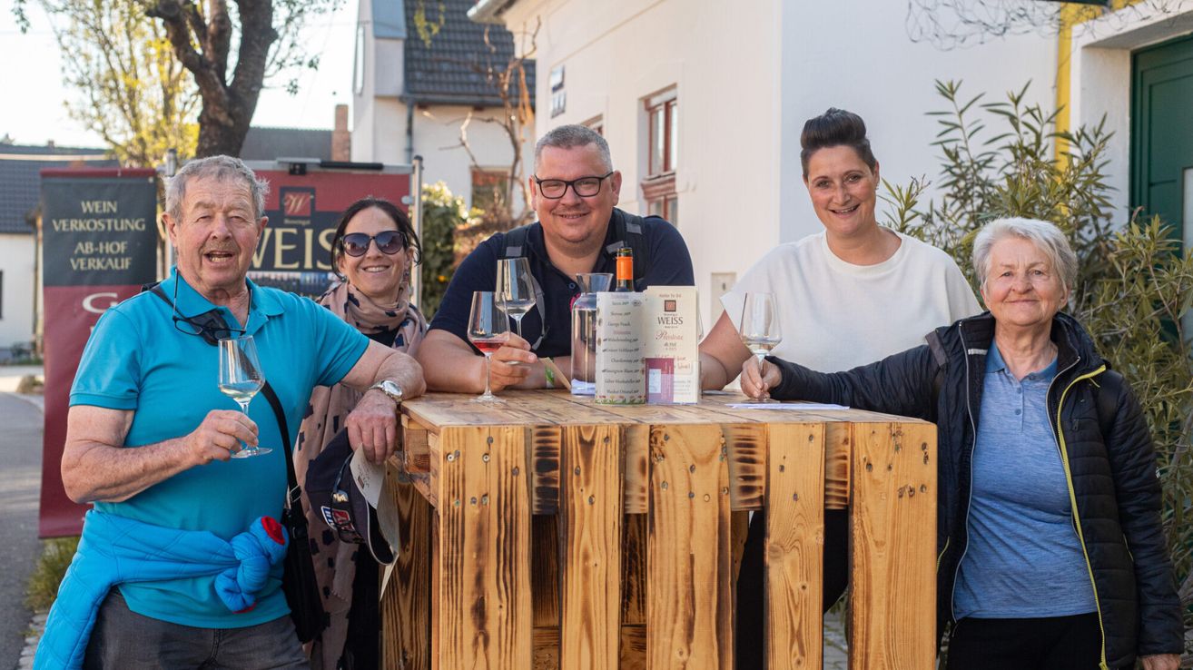 A group of four adults smiling, two men and two women, gather around a rustic wooden table with wine glasses, bottles, and a label reading 'Weiss'.