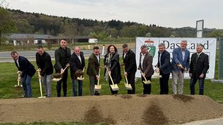 A group of people are holding shovels, standing in a line, and are probably participating in a groundbreaking ceremony. Behind them is a banner with the text Das Land.