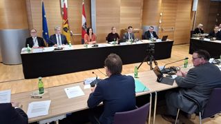 A conference room with a table, flags, and several people in suits. Four people are seated at the table with laptops and microphones. One person is sitting at a separate table.